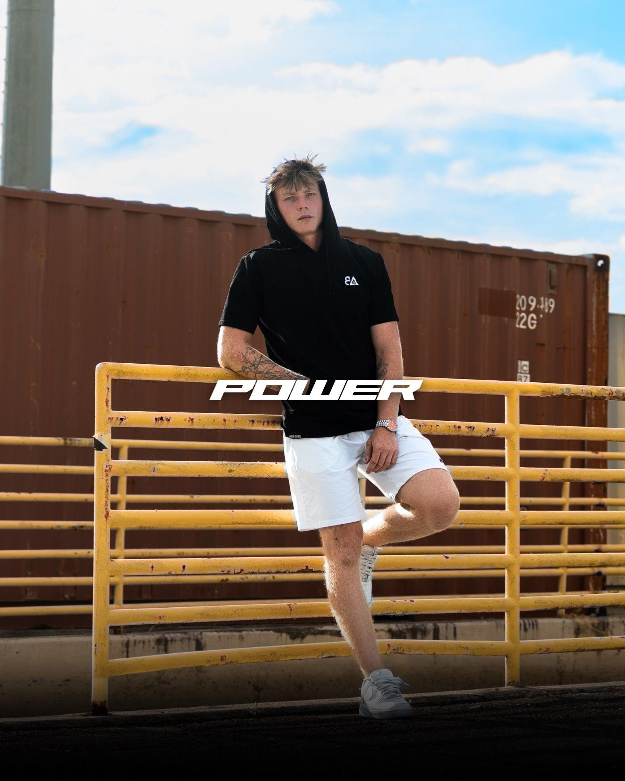 Person wearing a black hoodie with 'POWER' text, standing by a yellow metal gate with shipping containers in the background.