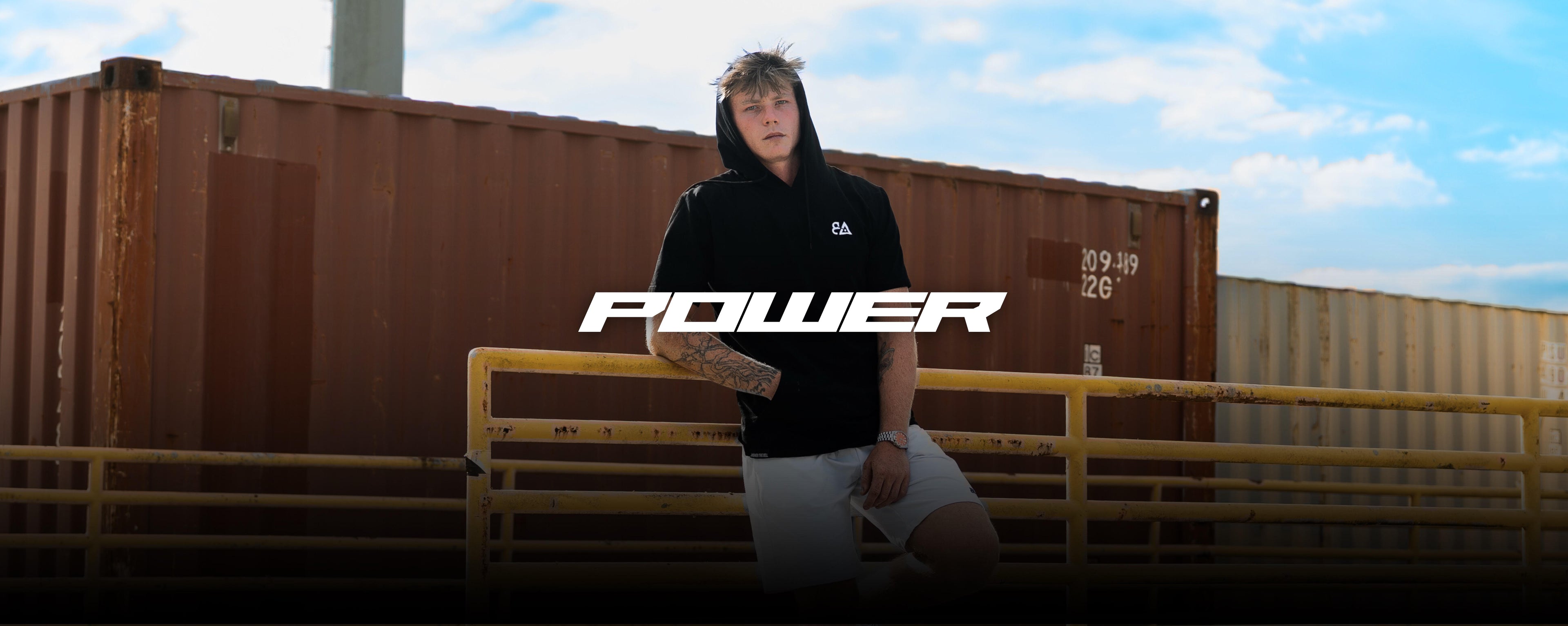 Person wearing a black hoodie with 'POWER' text, standing by a yellow metal gate with shipping containers in the background.1621243260e1af0c20-1